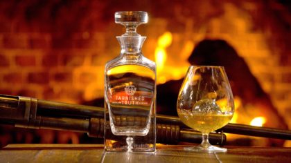 Tarnished Truth whiskey bottle and glass by the fire