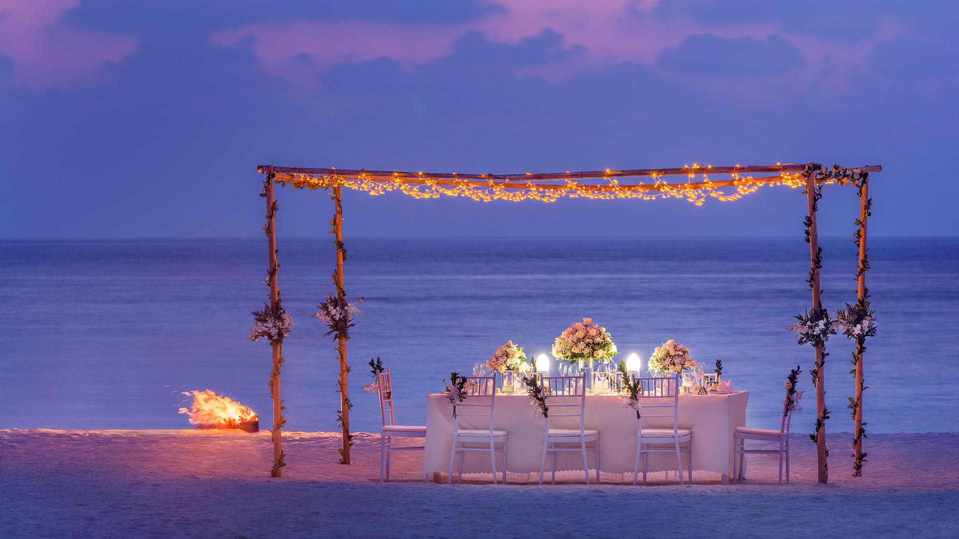 Canopy with lights over a fancy white table setting on the beach during dusk.