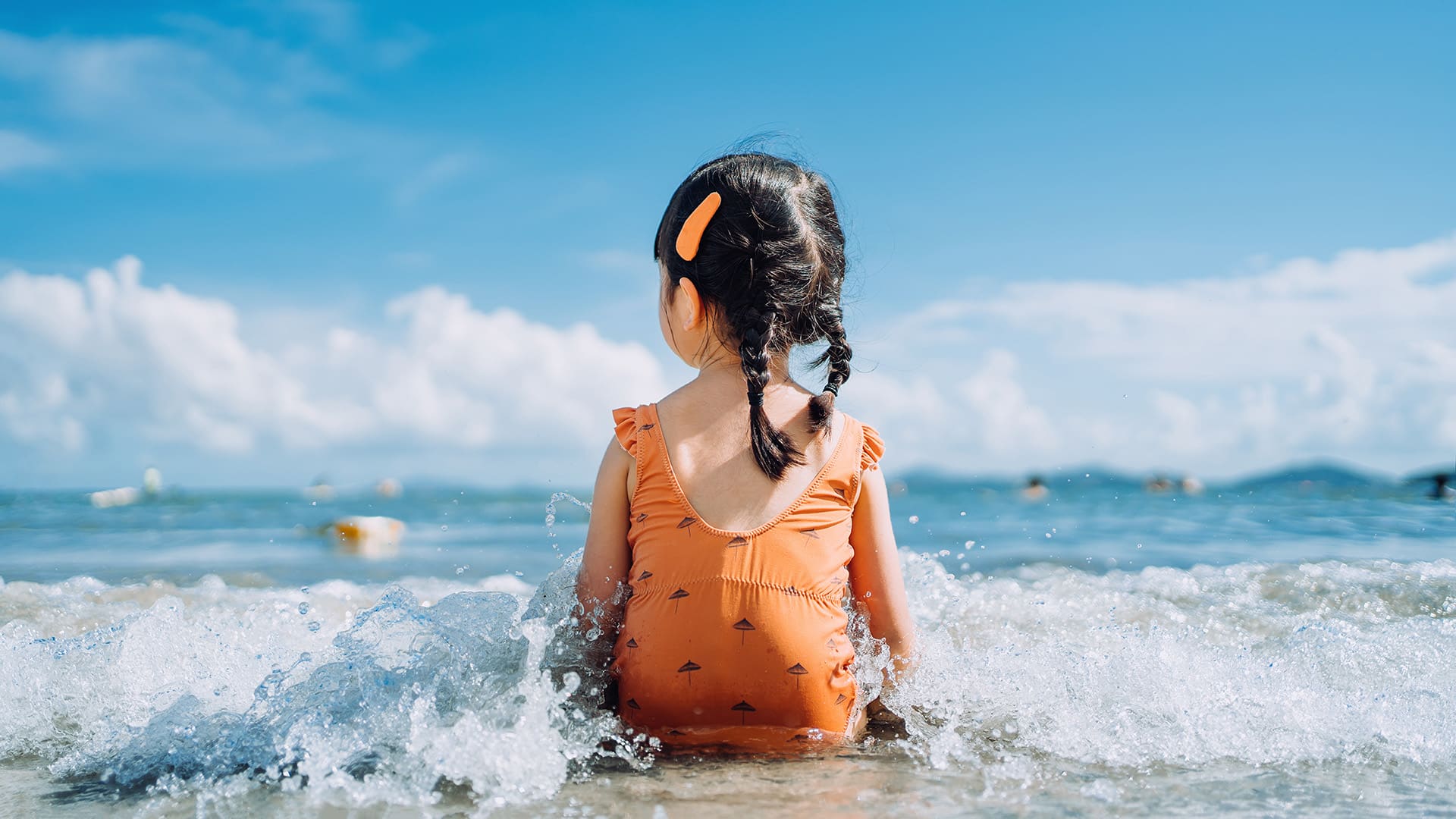 Toddler in orange bathing suit sitting on the beach in the waves.