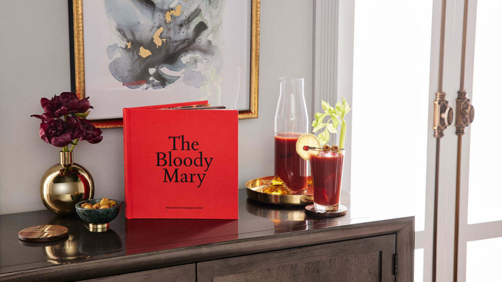 a copy of The Bloody Mary sits upright next to a carafe and full glass of Bloody Mary