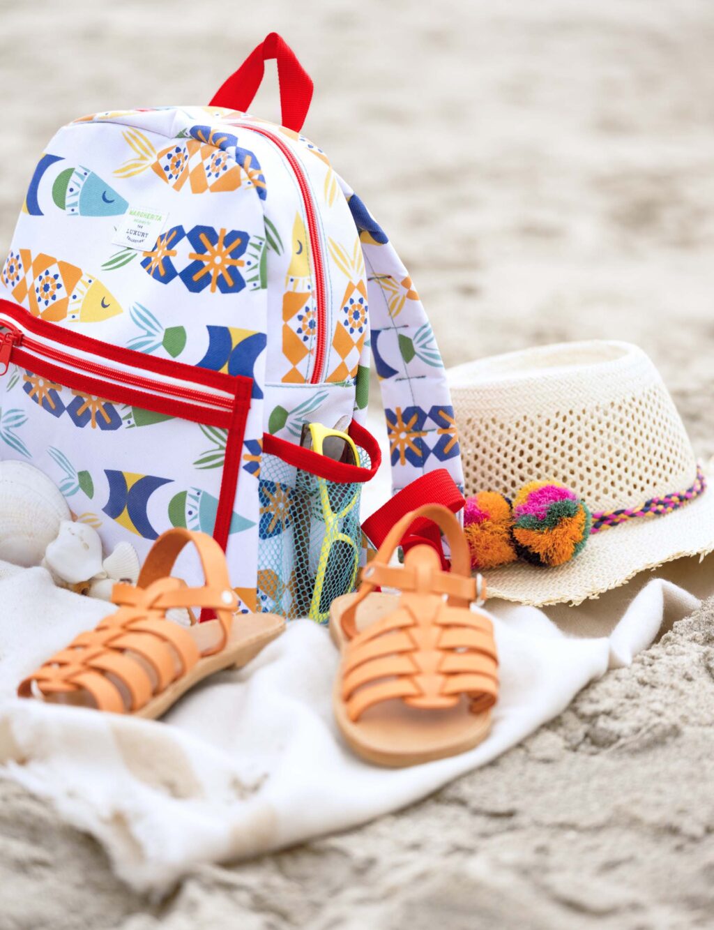 the Margherita Maccapani Missoni Children's Backpack for The Luxury Collection sits on a beach towel in the sand, along with children's sandles and a sun hat