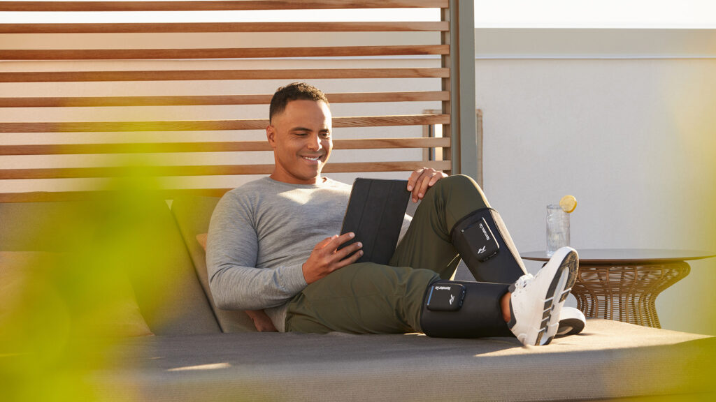 man sits on an outdoor couch; he reads on a tablet and has the Hyperice Normatec Go sleeves wrapped around his calves