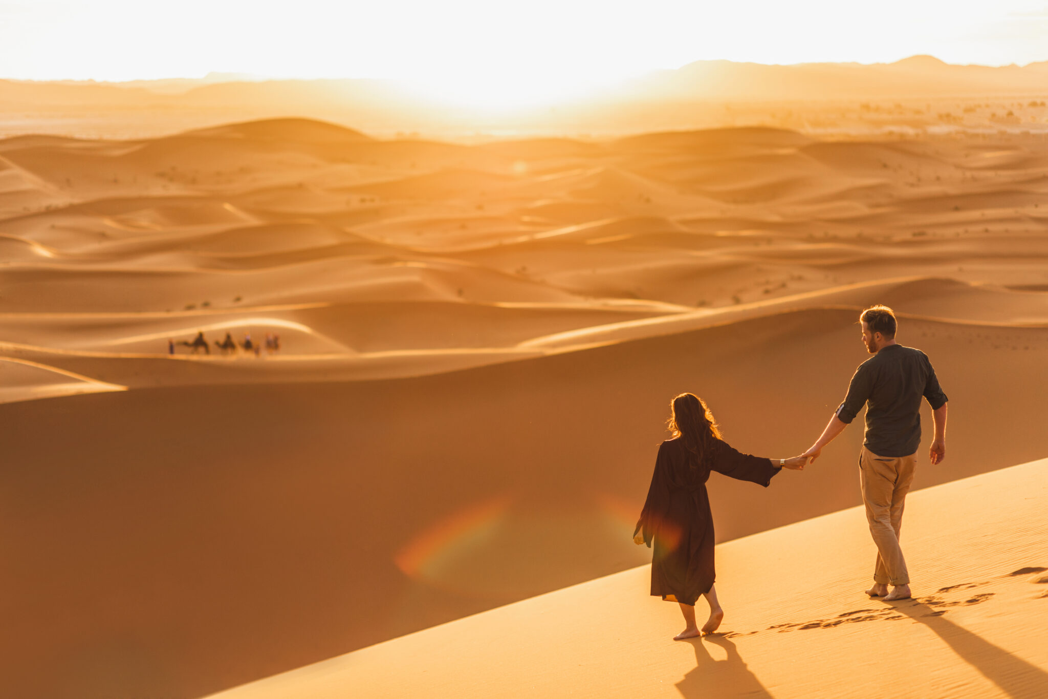 a woman and man hold hands and walk in the sand of a desert dune