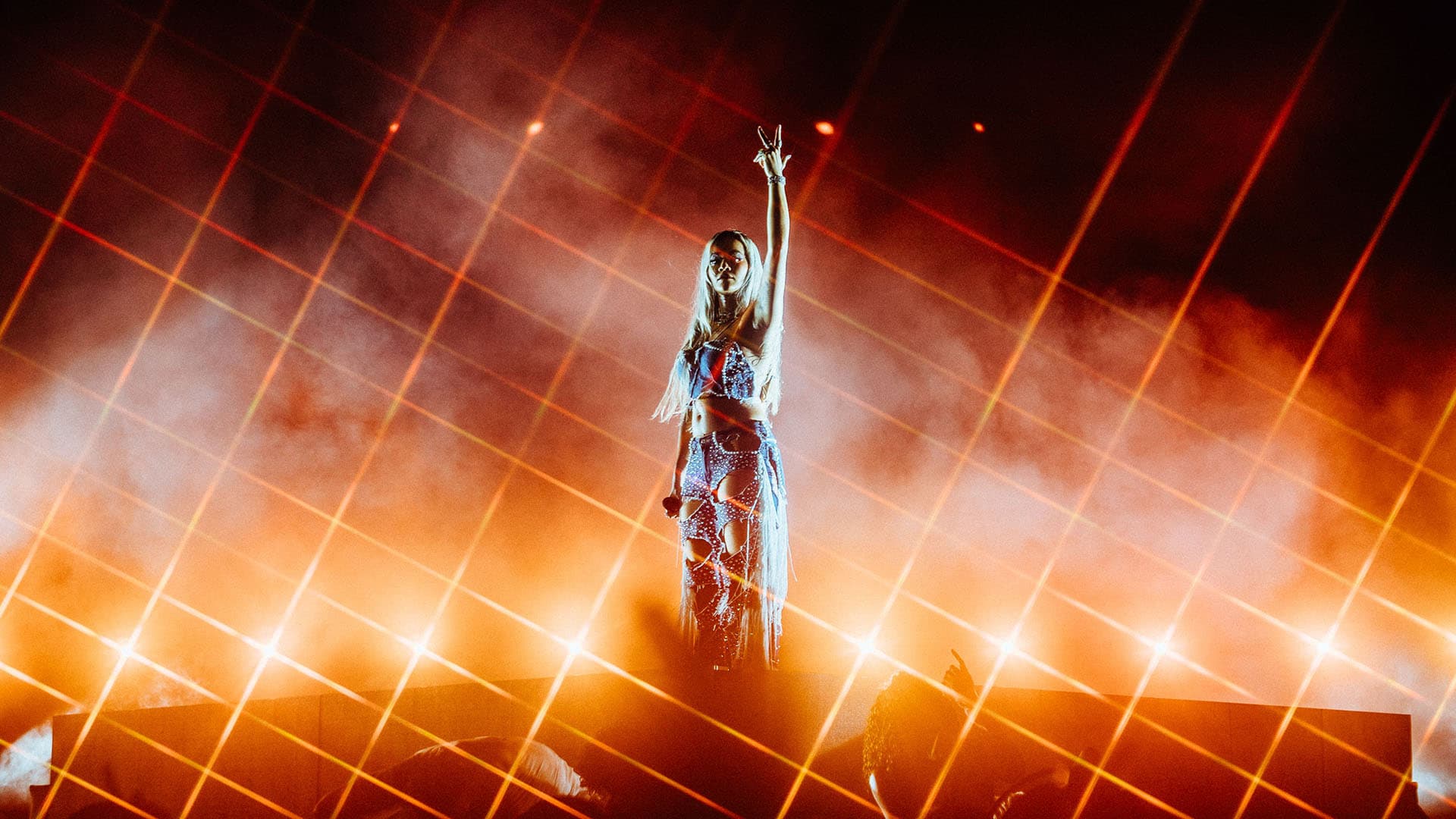 Rita Ora stands with one arm in the air on a stage with bright orange, grid-like lights