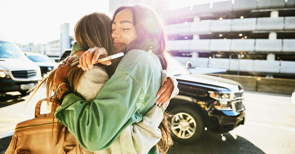 two women hug with cars passing by, at what appears to be the drop-off point an airport