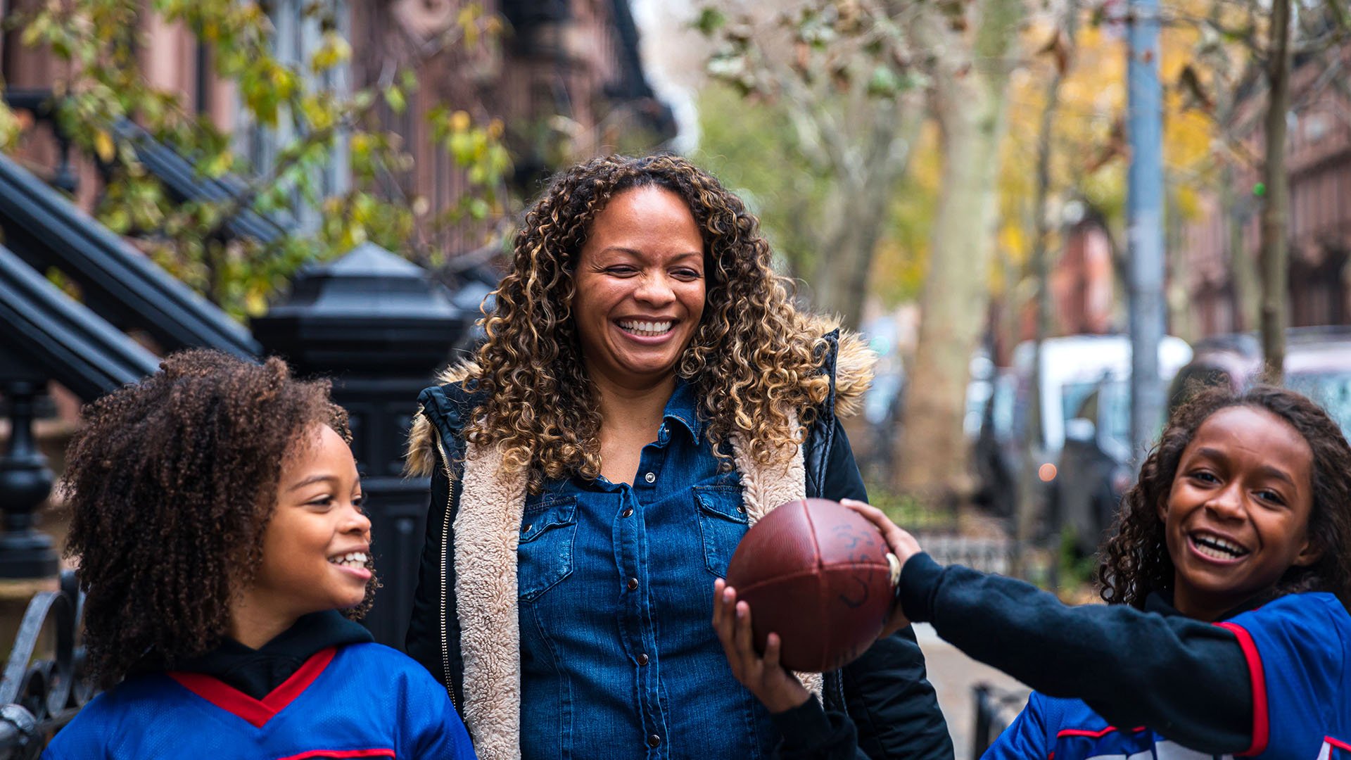 A mom and two daughters walk down a street in New York City; one is holding a football