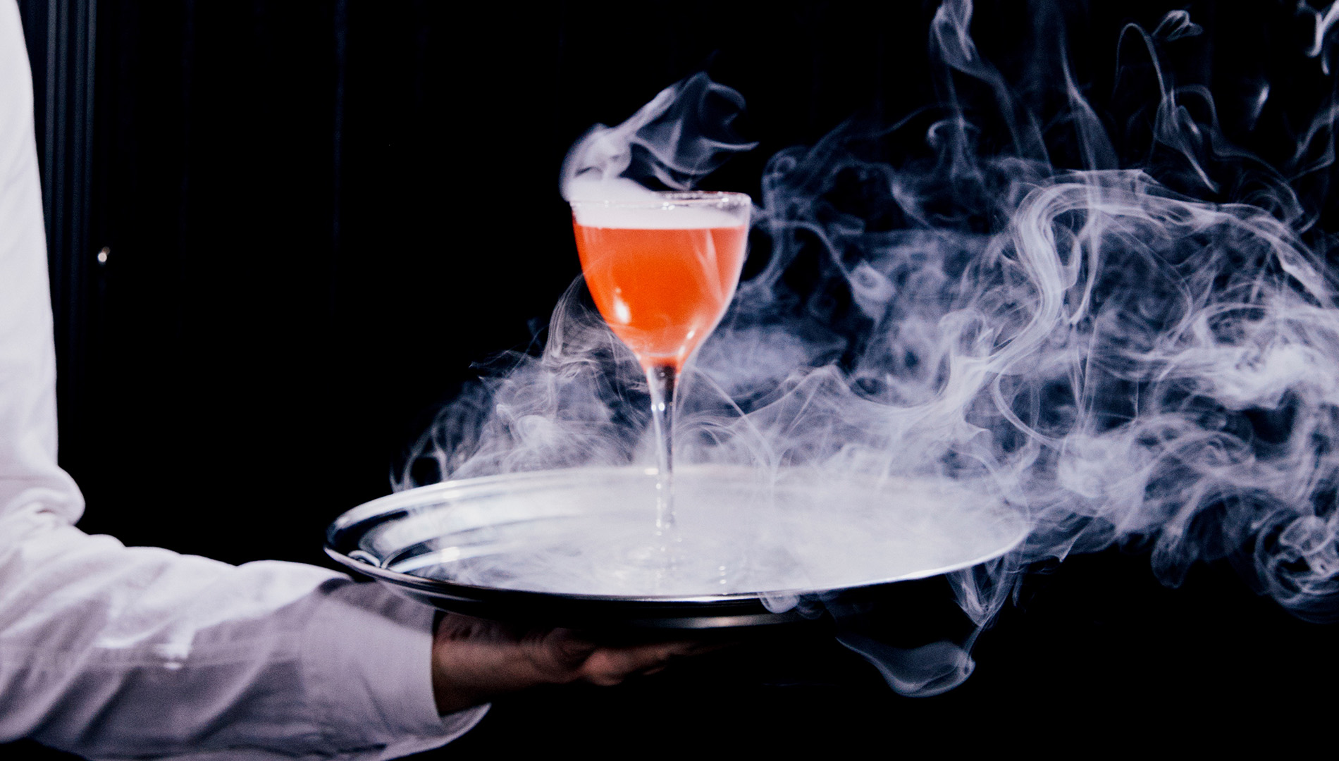 A waiter carries a fancy cocktail on a tray; smoke surrounds the glass