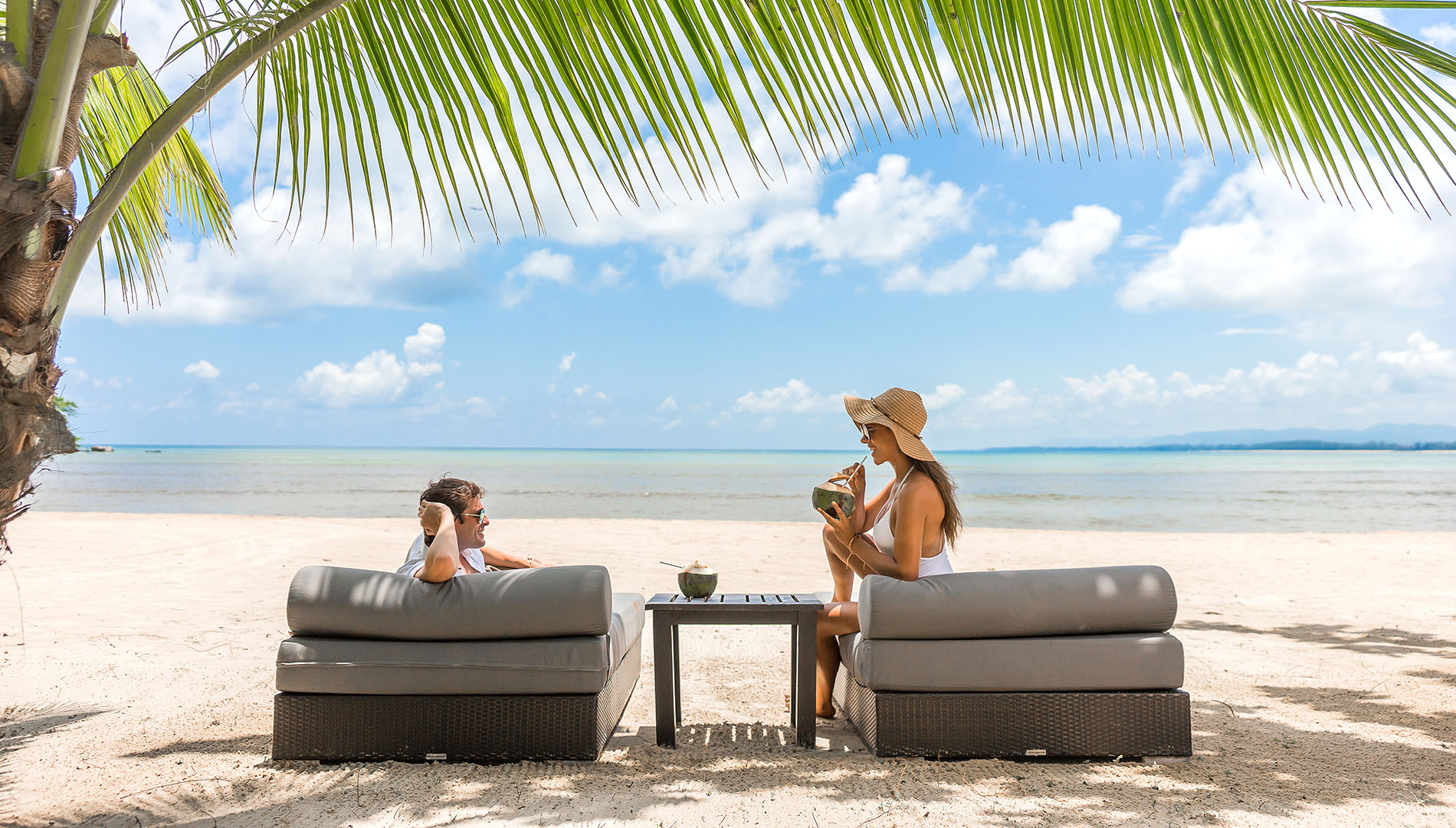 A man and woman sit on two plush chairs on a beach