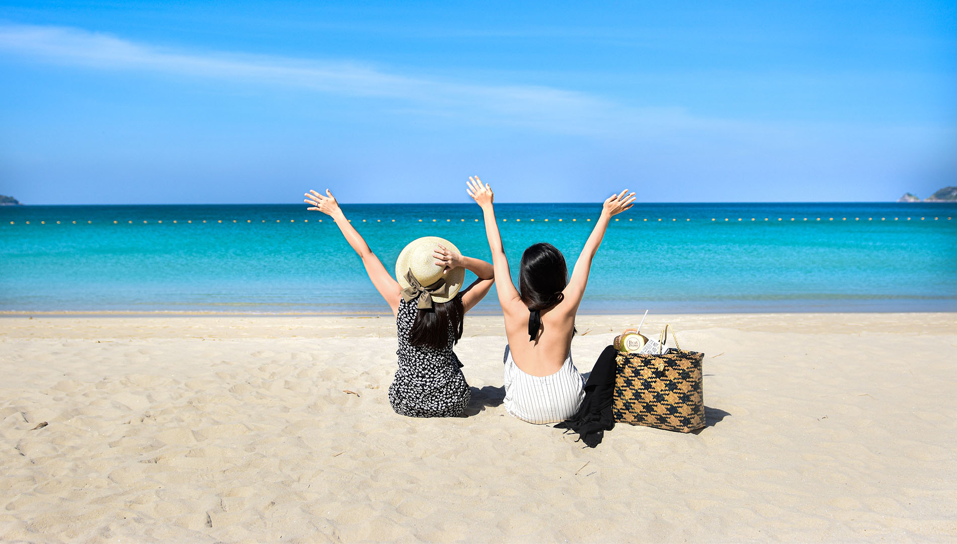 Two women sit on the beach with their arms raised