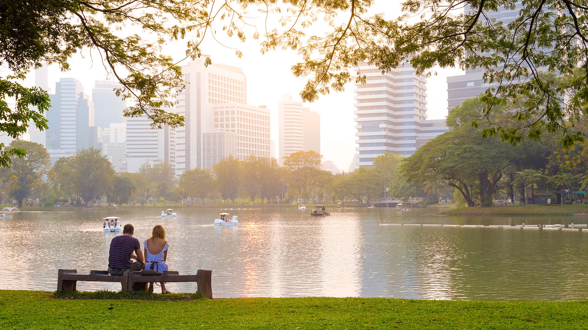 Two people sit on a bench in front of a lake in Lumpini Park in Bangkok, Thailand