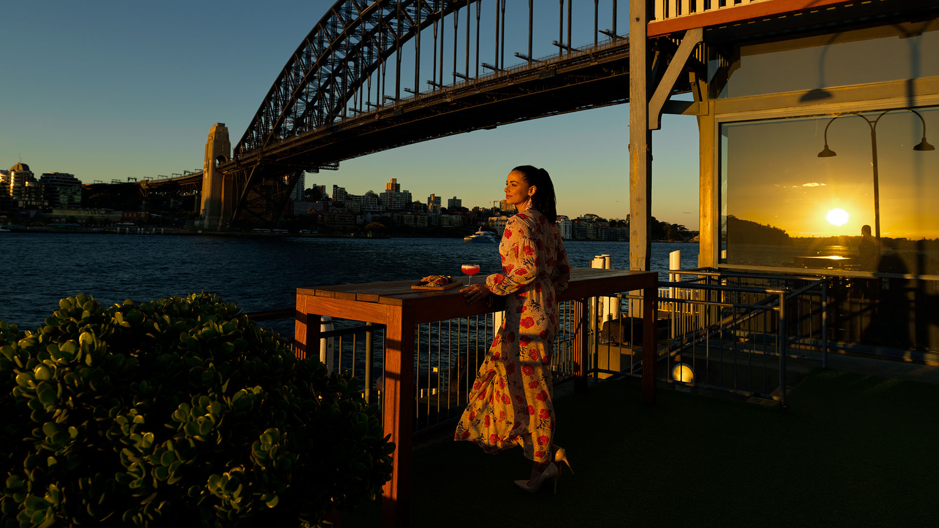 At sunset, a woman with a cocktail stands with the Sydney Harbour Bridge in the background