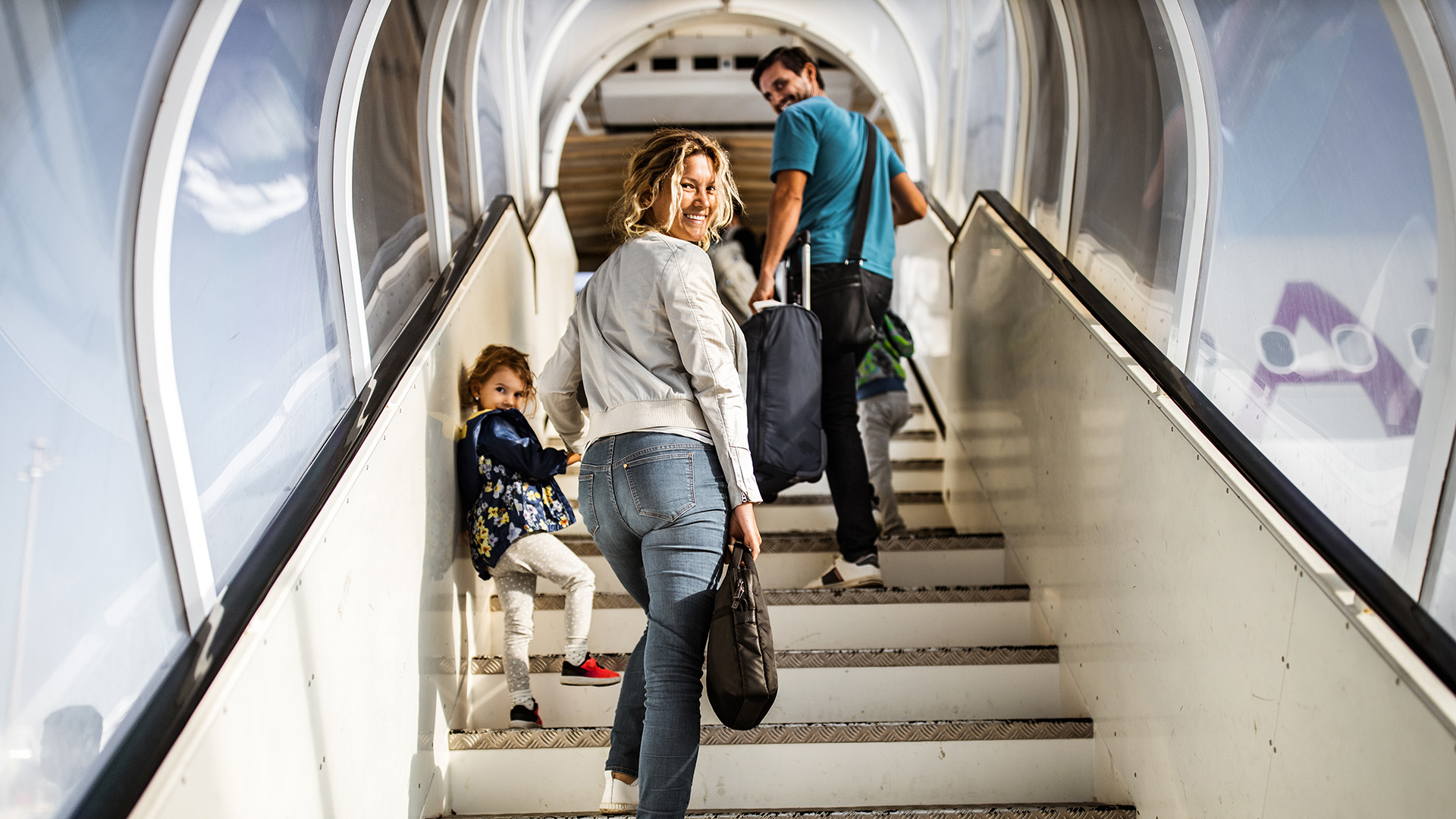 Two adults and a child looks back and smile as they ascend aircraft stairs up to a plane
