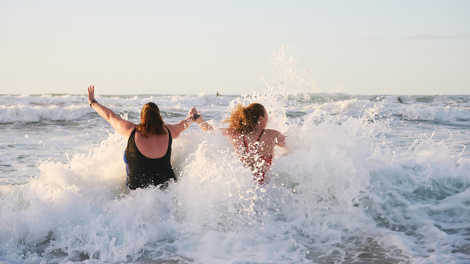 Two women hold hands as a wave crashes into them on a beach